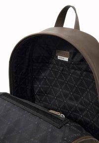 Mochila de cuero marrón con cierre de cremallera, que presenta un interior negro con patrón y un bolsillo de malla para organización. Marca sutilmente visible.