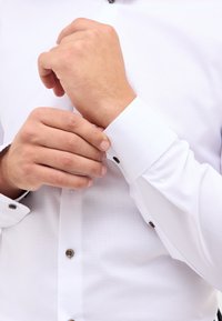 Camicetta bianca con finitura testurizzata, caratterizzata da un colletto senza bottoni, accenti a contrasto sui bottoni e un dettaglio di polsini ordinati.
