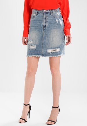 Kvinde iført en slidtdenim nederdel, rød langærmet top og sorte højhælede sandaler, stående mod en hvid baggrund.