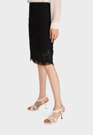 Donna che indossa una gonna nera di pizzo fino al ginocchio e sandali bianchi con tacco alto e cinturini, in piedi davanti a uno sfondo semplice.