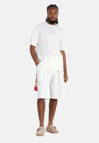 T-shirt blanc à col rond avec un petit texte rouge, associé à un short blanc jusqu'aux genoux avec un panneau latéral à motifs en beige et accents rouges.