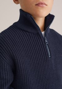 Mörkblå stickad tröja med hög krage och halvdragkedja. Har ett texturerat mönster och kontrasterande dragkedjedetaljer.