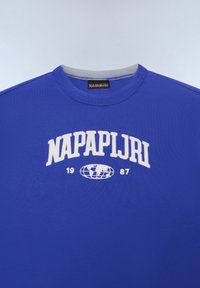 Mėlyna medvilninė marškinėlių su apvalia kaklo iškirpte, ant kurios pavaizduotas baltas "NAPAPIJRI" logotipas, pasaulio grafika ir metų "1987" užrašas priekyje.
