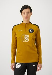 Long-sleeve mustard jersey with a quarter-zip, featuring black accents, a lion emblem, and a logo on the chest. Black pants with logo.