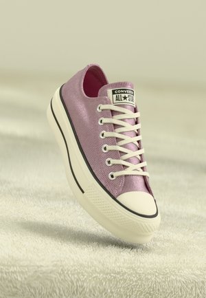 Roze Converse All Star sneaker met witte veters en zool, schuin geplaatst op een licht gestructureerd oppervlak.