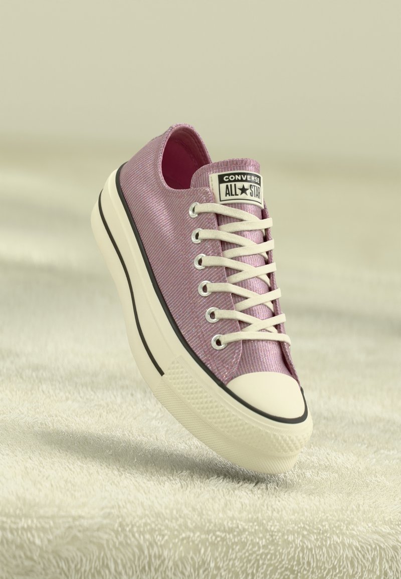 Baskets Converse All Star roses avec lacets et semelle blancs, inclinées sur une surface claire et texturée.