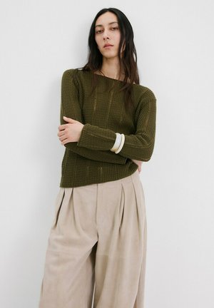 Woman with long dark hair wears an olive green knit sweater, beige pleated pants, white bangles, standing with arms crossed against a white wall.