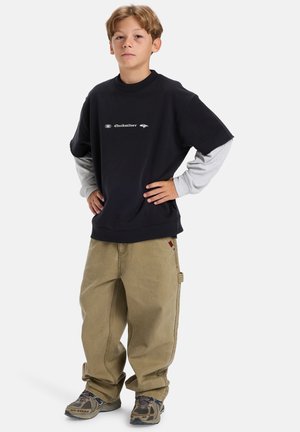 Joven de pie con las manos en las caderas, vistiendo capas de manga larga negra y gris, pantalones anchos color beige y zapatillas marrones, sobre fondo blanco.