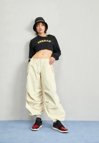 Mladý člověk se na šedé zdi, oblečený v černé zkrácené mikině "Jordan", béžových volných kalhotách, černo-červených teniskách a černém klobouku typu bucket hat.