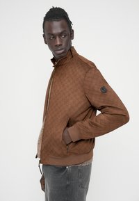 Καφέ μπουφάν τύπου bomber με υφασμάτινο μοτίβο, κλείσιμο με φερμουάρ, γιακά όρθιο και πλαϊνές τσέπες. Διαθέτει λογότυπο στο μανίκι αριστερά.