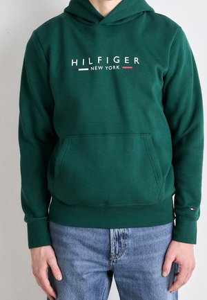 Osoba ubrana w ciemnozieloną bluzę z kapturem z napisem „HILFIGER NEW YORK” z przodu oraz jasnoniebieskie dżinsy, stojąca na jednolitym tle.