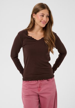 Jeune femme aux longs cheveux bruns porte un haut côtelé marron foncé à manches longues et un pantalon rose, debout avec les mains derrière le dos, souriante.