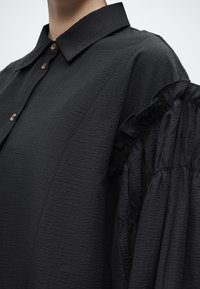 Μαύρο πουκάμισο με υφή, με δομημένο γιακά και φουσκωτά, ρυγχωτά μανίκια που φαίνονται από τον ώμο μέχρι το μέσο του κορμού.