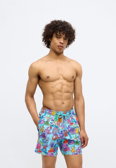 Tânăr cu păr creț, îmbrăcat în pantaloni scurți de baie colorați, de un albastru vibrant, cu un model de corali și pești subacvatici, stând pe un fundal alb.