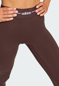 Brown leggings with a seamless design, featuring a white "adidas" logo on the waistband and subtle texture throughout the fabric.