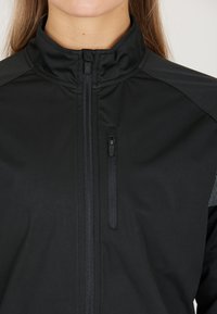 Schwarze Sportjacke mit hohem Kragen, Reißverschluss vorne und einer kleinen Reißverschlusstasche. Strukturiertem Stoff mit glatter Oberfläche und reflektierenden Akzenten.