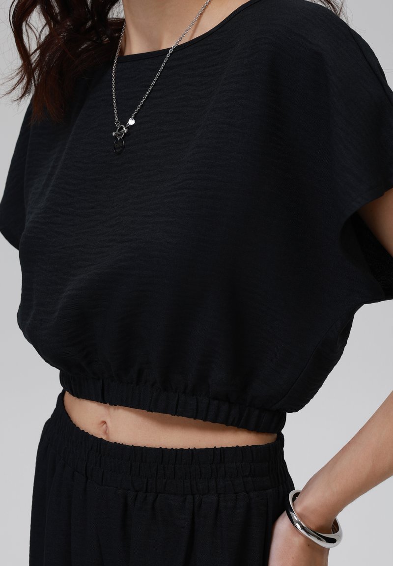 Schwarzes Cropped Top aus strukturiertem Stoff, mit weiten kurzen Ärmeln und einem elastischen Saum. Kombiniert mit passenden hochtaillierten Hosen und einem silbernen Armband.