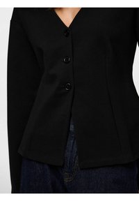 Juoda ilgomis rankovėmis palaidinė su V formos iškirpte, priekyje penki juodi mygtukai ir aptemptas siluetas, derinama su tamsiais džinsais.