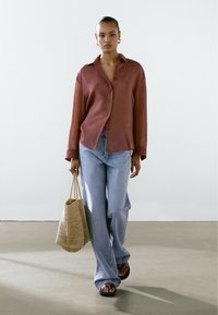 Rustrød linned skjorte med knapper, lyseblå brede jeans, vævet beige håndtaske og sorte sandaler, mod en neutral baggrund.