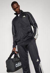 Melnas sporta komplekts ar baltām svītrām un Adidas logo. Modelis tur melnu sporta somu. Audums izskatās gluds un viegls.