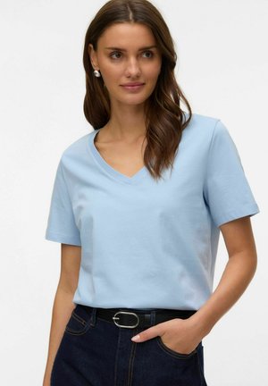 Woman with long brown hair wearing light blue V-neck T-shirt and dark jeans with black belt, hand in pocket, looking slightly right.