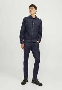Chaqueta de mezclilla oscura con dos bolsillos en el pecho y cierre de botones, combinada con pantalones de mezclilla oscura a juego y zapatos negros de cordones.