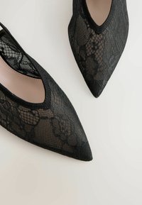 Čierne čipkované lodičky na vysokom opätku so špicatou špičkou, s prepracovanými kvetinovými vzormi a elegantným dizajnom, doplnené o hladké vnútorné podšívku.