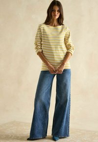 Gestreifte gelbe und weiße Langarmbluse, blaue weite Jeans und türkisfarbene Schuhe. Weicher Stoff mit lässigem Schnitt. Neutraler Hintergrund.