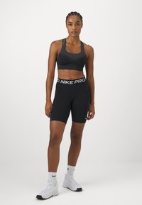 Crna sportska grudnjak s teksturiranim materijalom, u kombinaciji s crnim Nike Pro kratkim hlačama s bijelim logotipom i odgovarajućim bijelim sportskim cipelama.