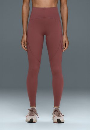 Personne debout portant des leggings bordeaux taille haute et des chaussures de course rose clair avec des chaussettes grises sur un fond gris uni.