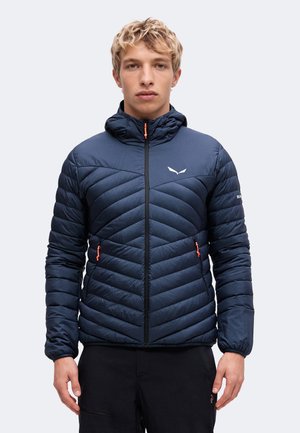 Jonge man die een navyblauwe doorgestikte jas met capuchon draagt, met een rits aan de voorkant en oranje ritstrekkers, staand tegen een effen lichte achtergrond.
