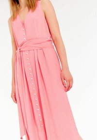Robe maxi sans manches rose avec une taille à nouer, dotée d'un décolleté en V et d'accents de boutons blancs courant le long de l'avant. Texture de tissu lisse.