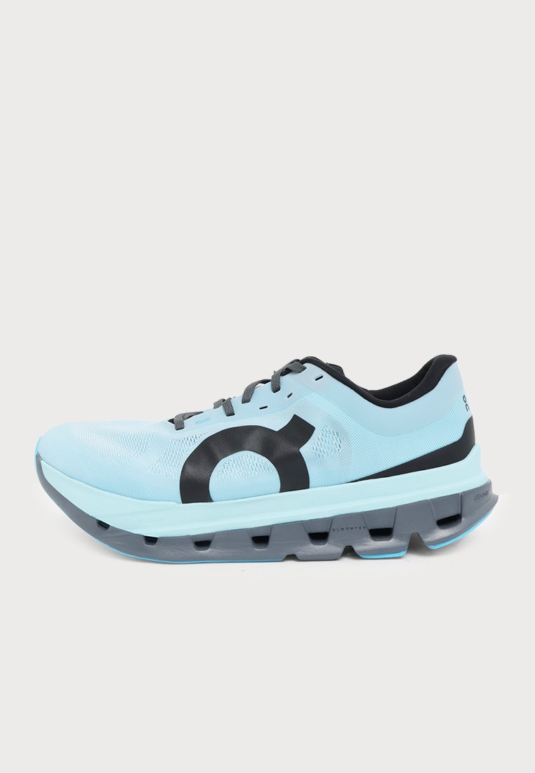 Zapatilla de running azul claro con cordones negros y logotipo, una suela gruesa gris segmentada, vista de perfil.