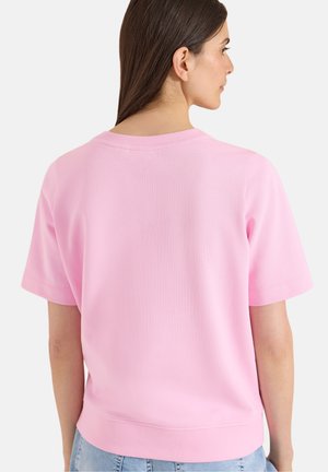 Femme aux longs cheveux bruns portant un t-shirt uni rose clair à manches courtes et un jean bleu clair, vue de dos.