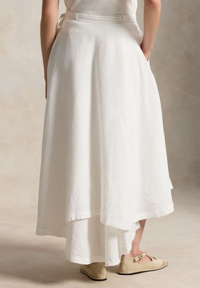 Moteris dėvi ilgą, sluoksniuotą baltą sijoną ir smėlio spalvos butas batus, stovinti su rankomis kišenėse prieš neutralią foną.