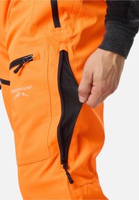 Heldere oranje broek met zwarte ritszakken en mesh voering. Bevat een logo en dubbelzijdige stof voor verbeterde duurzaamheid.