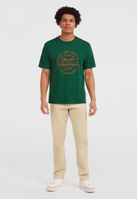 Grøn bomuldst-shirt med orange grafisk tekst og bjergmotiver, parret med beige bukser og hvide sneakers.