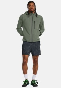 Chaqueta verde con capucha, cierre frontal y bolsillo en el pecho, combinada con pantalones cortos negros con estampado, calcetines blancos y zapatos negros con acentos en neón.