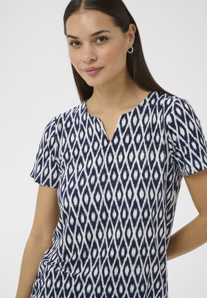 Frau trägt ein kurzärmeliges, marineblau-weißes Oberteil mit geometrischem Muster und V-förmigem Ausschnitt, lächelt leicht vor einem schlichten Hintergrund.