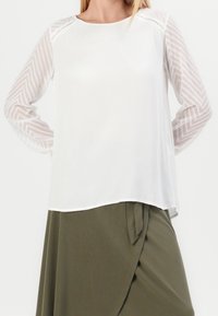 Haut blanc avec des manches longues en voile à motifs, arborant des designs en chevron. Le corps est lisse et de coupe décontractée, assorti à une jupe unie olive.