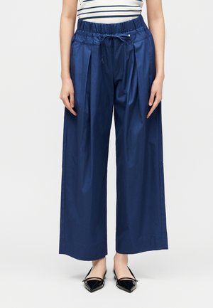 Femme portant un pantalon bleu marine taille haute, jambes larges avec un cordon à la taille, associé à des ballerines noires pointues et un haut rayé.