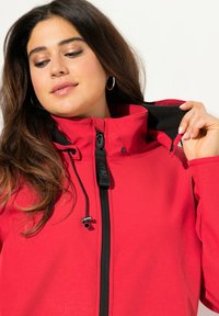 Giacca rossa con colletto alto, chiusura con zip, accenti neri, coulisse regolabile e tessuto testurizzato. Modello che tiene il colletto.