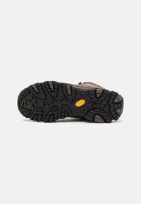 Podeszwa buta trekkingowego z czarną gumową podeszwą, z głębokimi wypustkami zapewniającymi przyczepność oraz logo Vibram w pomarańczowym heksagonie.