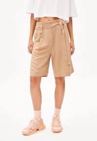 Beige tailored shorts with a high waist, tie belt, and side pockets, paired with light peach sneakers and ribbed socks. Smooth texture.