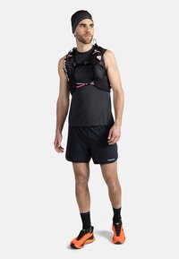 Černá běžecká vesta s nastavitelnými popruhy, vyrobena z lehké látky, v kombinaci s černým bezrukávovým tričkem a šortkami. Jasně oranžové běžecké boty.