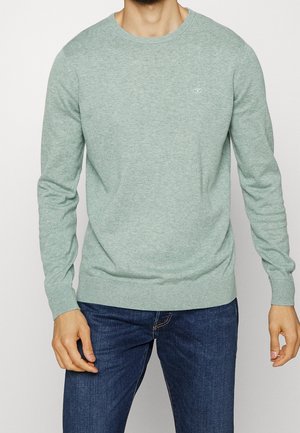 Man draagt een lichtgroene trui met ronde hals en geribbelde boorden en zoom, gecombineerd met donkerblauwe jeans tegen een effen achtergrond.