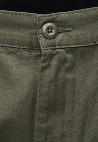 Ύφασμα ελαιόχρωμο με υφή, κλείσιμο με μεταλλικό κουμπί και εμφανείς ραφές, με λείο ζωνάρι και λεπτομέρεια ραφής στο μπροστινό πάνελ.