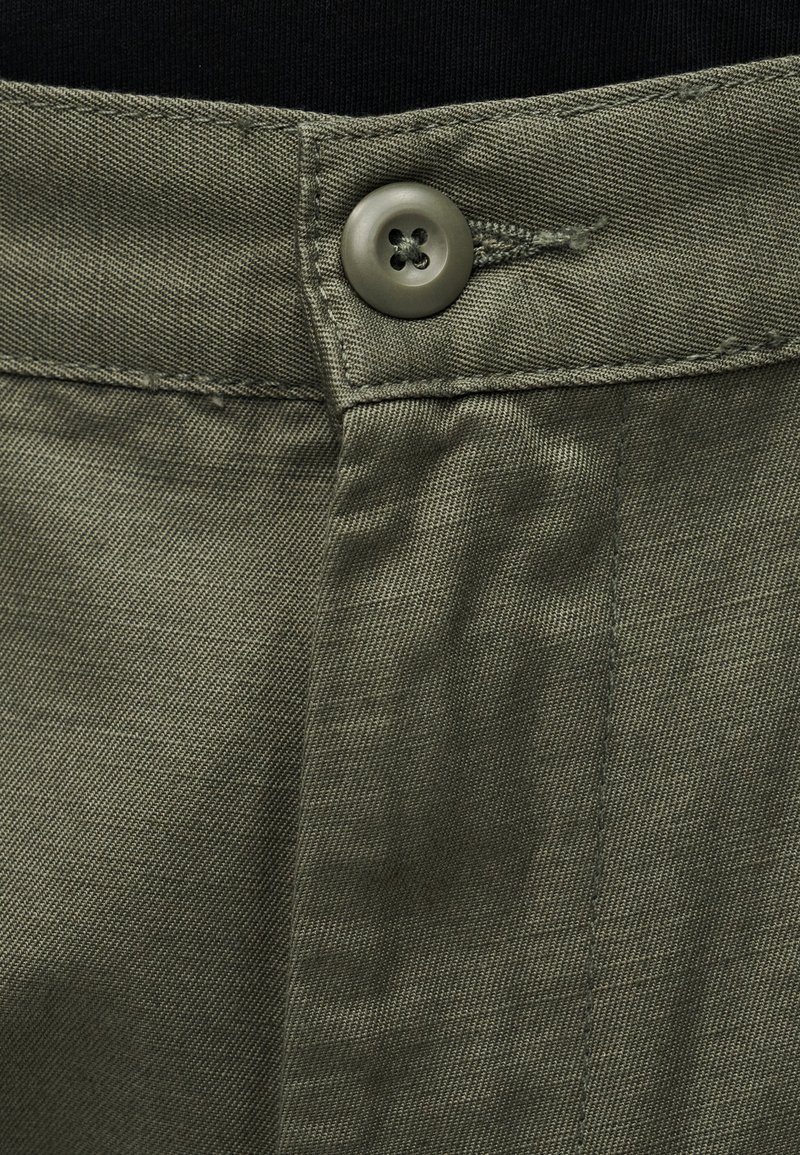 Tissu texturé vert olive avec une fermeture à bouton métallique et des coutures visibles, présentant une ceinture lisse et un détail de couture sur le panneau avant.