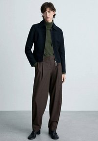 Giacca nera con zip, dolcevita verde, pantaloni marroni plissettati, scarpe nere eleganti. Tessuto fluido con un design strutturato, stile minimalista.