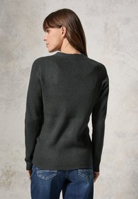 Dunkelgrüner, gerippter Pullover mit langen Ärmeln und rundem Ausschnitt, kombiniert mit blauen Jeans. Die Textur ist grob und gestrickt.